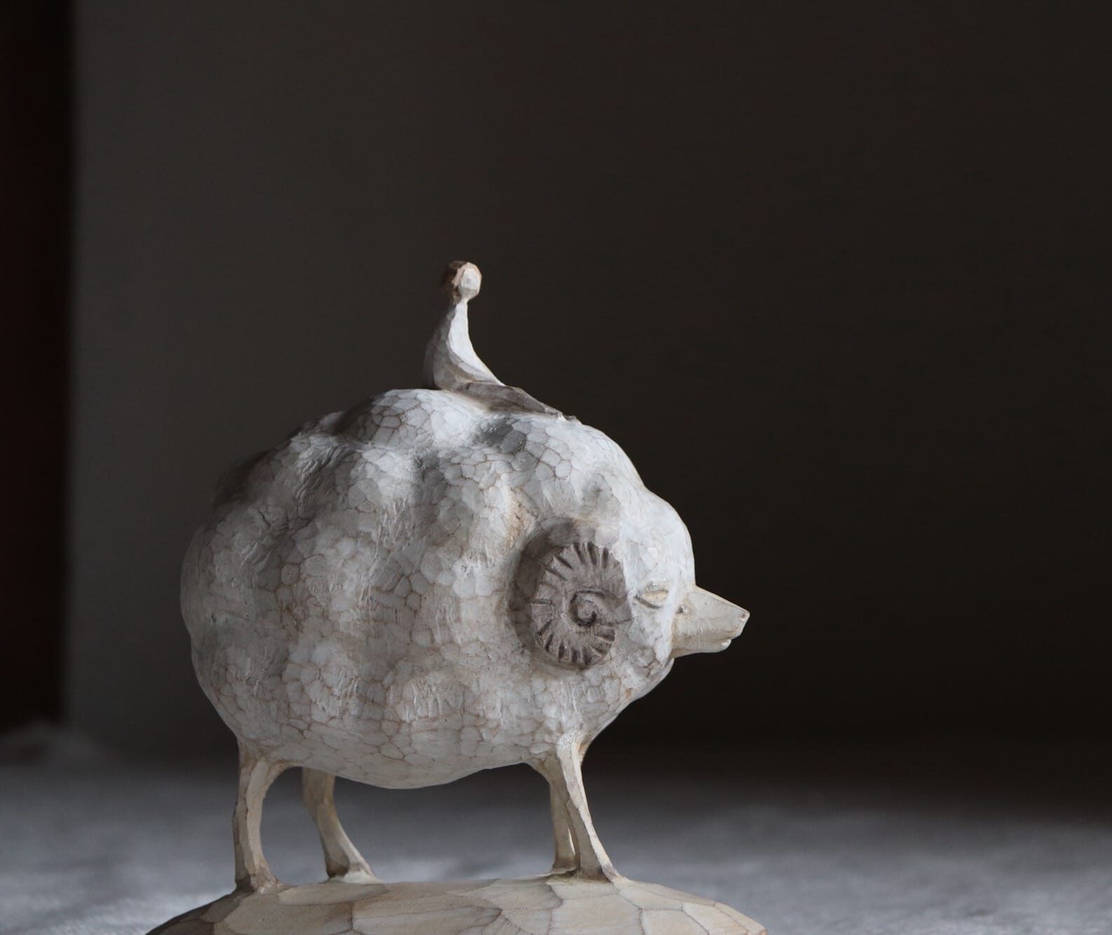 A photograph of a small wooden carved statue of a sheep, shown side-on against a black backdrop. The sheep’s puffed out coat is disproportionately large compared to its head and legs. A curled horn is visible against the side of the sheep’s head in amongst the woollen coat. On top of the sheep is a very small seated human figure.